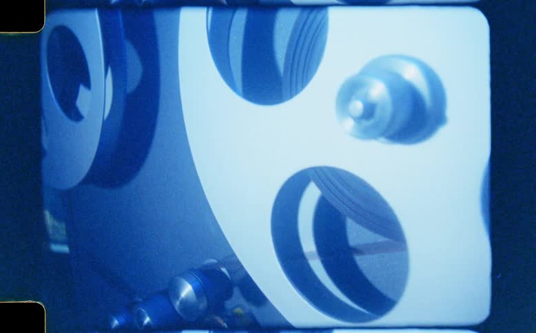 A vintage metal film reel with circular cutouts spins on a projector, captured on authentic 8mm film with a distinct blue tint