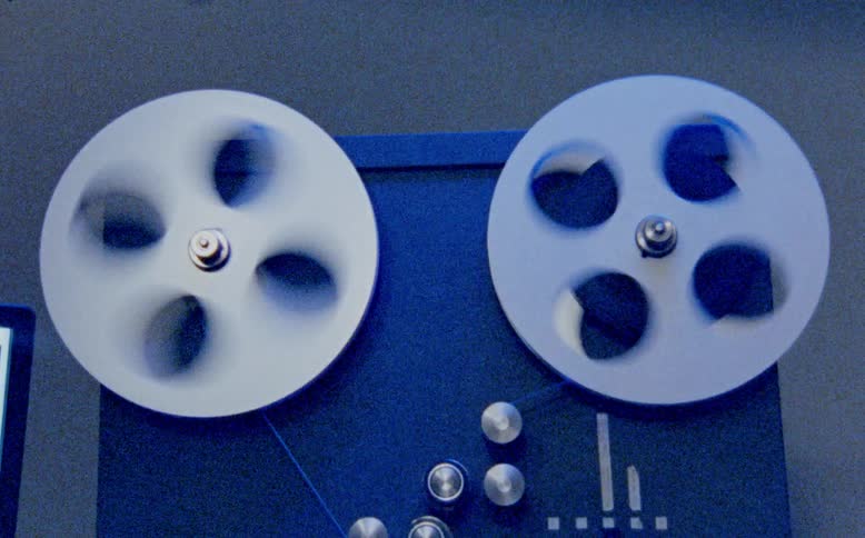 A vintage reel-to-reel tape recorder plays with fast spinning reels in a seamless loop with a grainy film look and changing light