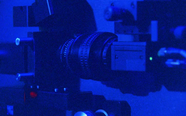 A grainy, blue-lit montage shows a close-up of a laser machine and then two classic film reels