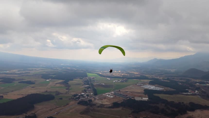 A bright green paraglider soars over a vast rural valley and mountain landscape under a cloudy sky