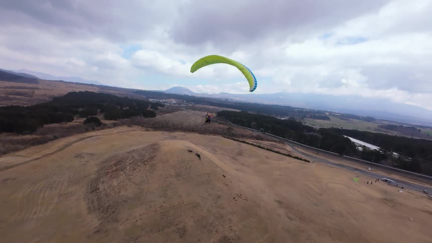 Dynamic aerial footage follows a lone paraglider as they soar over rural fields and hills on a cloudy day