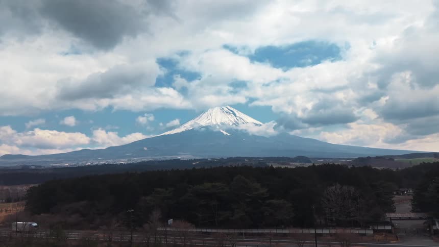 A majestic aerial pullback reveals the snow-capped peak of Mount Fuji towering over a vast forest and rural landscape under a dramatic cloudy sky
