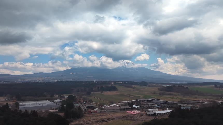 An epic aerial view of a snow-capped mountain towering over a rural Japanese town under a dramatic cloudy sky