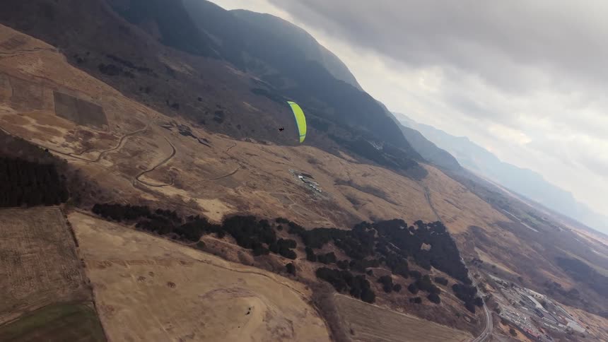 Dynamic aerial view follows a bright green paraglider soaring gracefully over a sprawling mountain valley on a cloudy day