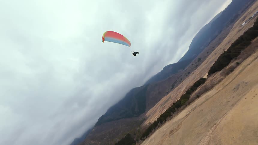 An FPV drone captures exhilarating aerial footage of a tandem paraglider soaring over the vast, rural landscape near a majestic mountain