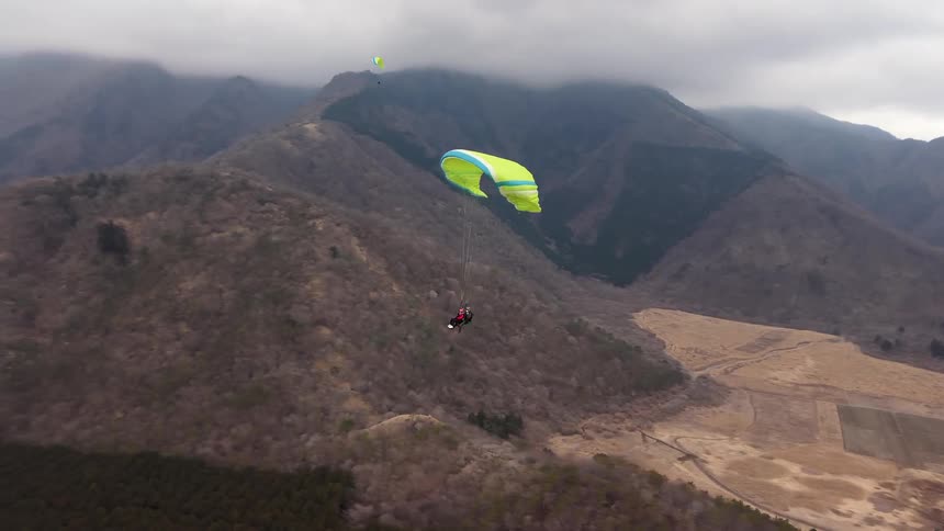 An aerial shot follows a lone paraglider soaring over a vast mountain valley on a cloudy day