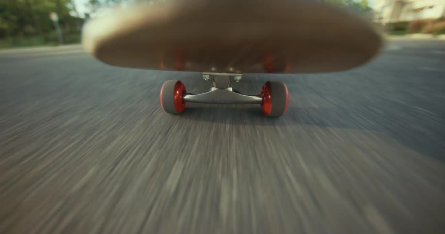 A low-angle point-of-view shot captures the back wheels of a skateboard spinning rapidly as it cruises down a sunlit street