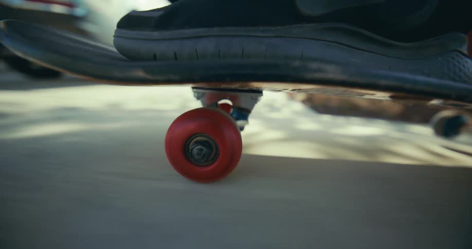 A close-up, low-angle shot shows a red skateboard wheel spinning as it rolls down a sunny sidewalk
