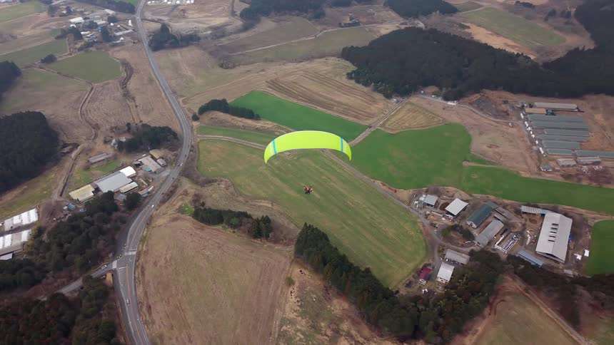 A bright green paraglider soars peacefully over a vast rural landscape of patchwork fields, forests, and small villages