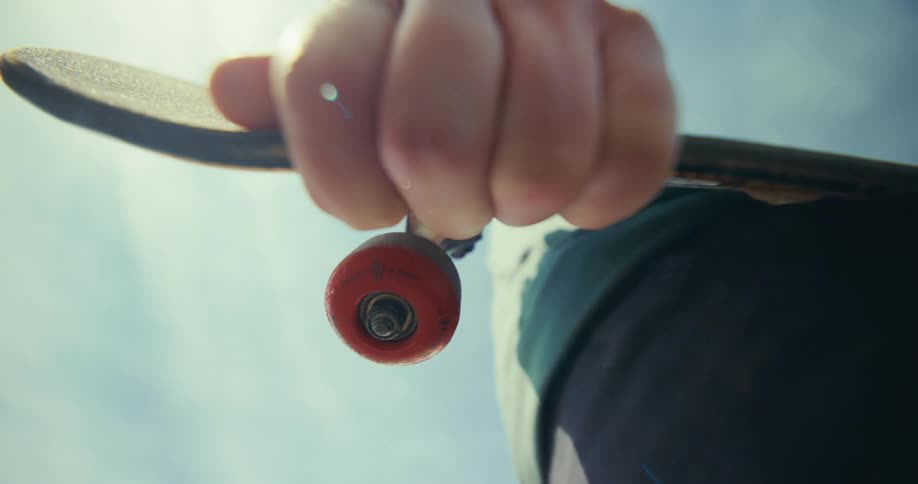 A worm's-eye view captures a person's hand gripping a skateboard with a red wheel against a bright, sunlit sky