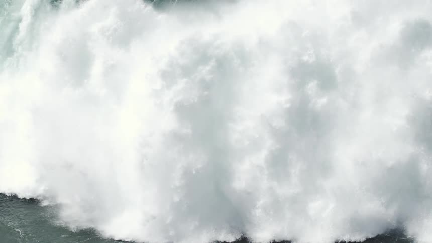 An epic slow-motion shot captures a lone surfer riding down the face of a massive, powerful ocean wave as it crashes