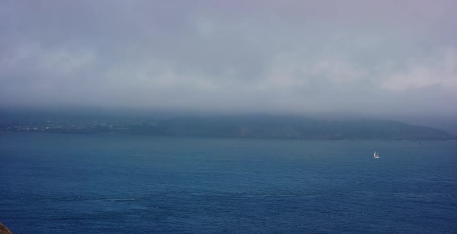 A solitary sailboat glides across the vast, misty ocean under a thick layer of fog