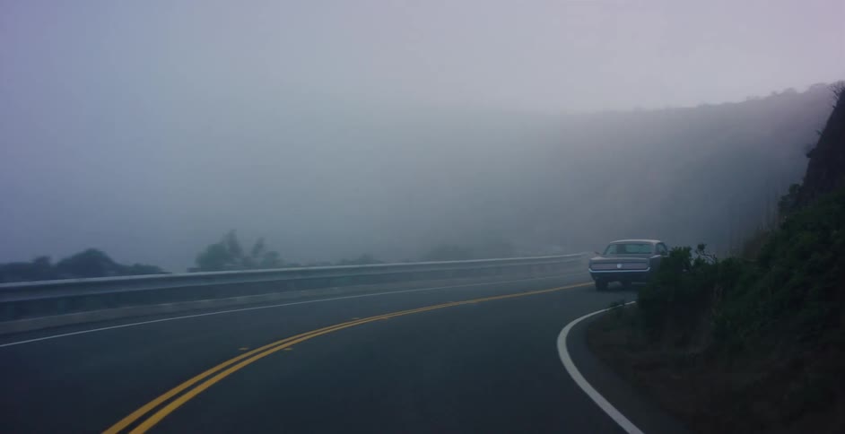 A vintage American muscle car slowly drives along a winding, foggy two-lane road next to a mountain