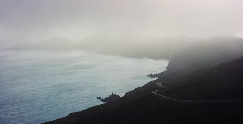 A winding road follows the rugged coastline, disappearing into the thick fog over the ocean