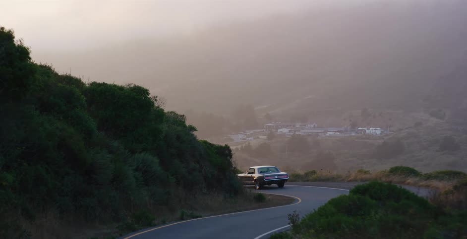 A vintage car drives along a winding mountain road through a hazy valley at dusk