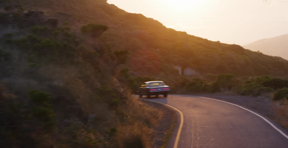 A classic black American muscle car drives around a scenic bend on a coastal highway as the golden sun sets
