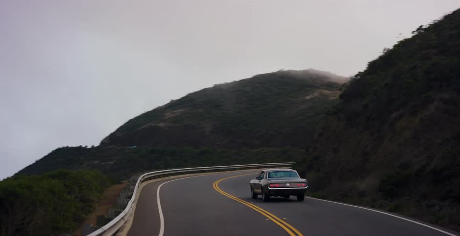 Rear view of a classic silver car navigating a winding road through foggy, green hills