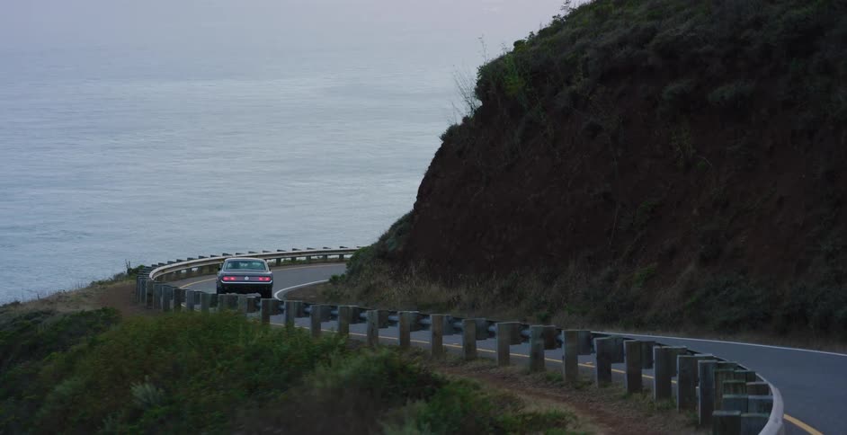 A classic muscle car navigates a winding coastal highway overlooking the vast ocean at twilight