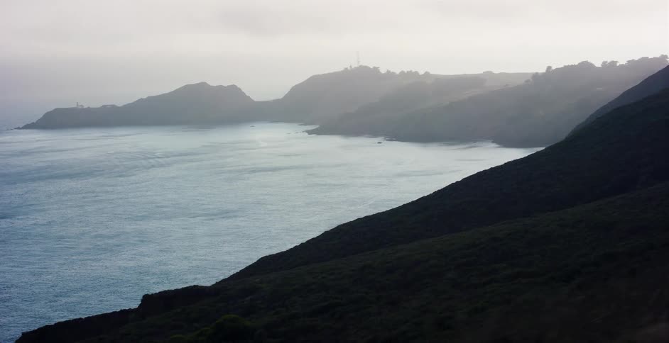 A misty aerial view reveals the foggy northern California coastline with dark rolling hills and calm ocean waters