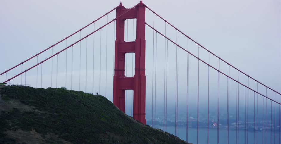 A lone person walks along a green hill with a close-up view of the Golden Gate Bridge tower shrouded in fog