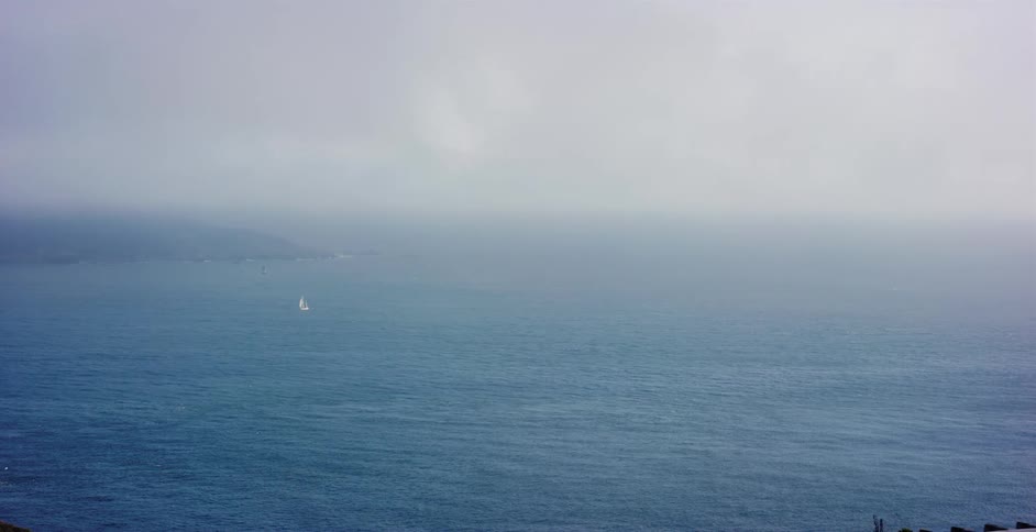 A solitary sailboat navigates the vast, foggy ocean on an overcast day
