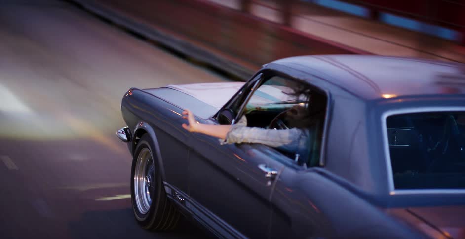A carefree woman drives a classic Ford Mustang at dusk, her arm out the window feeling the wind as her hair blows freely