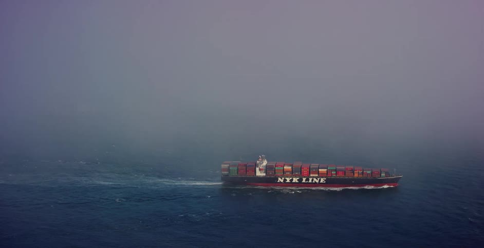 An aerial view captures a large container ship sailing through the vast open ocean, its journey shrouded in thick, mysterious fog
