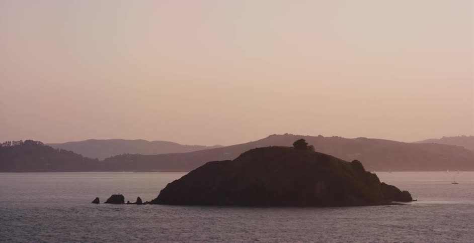 A serene wide shot captures the silhouette of a small island in a calm bay against a hazy, pastel sunset sky