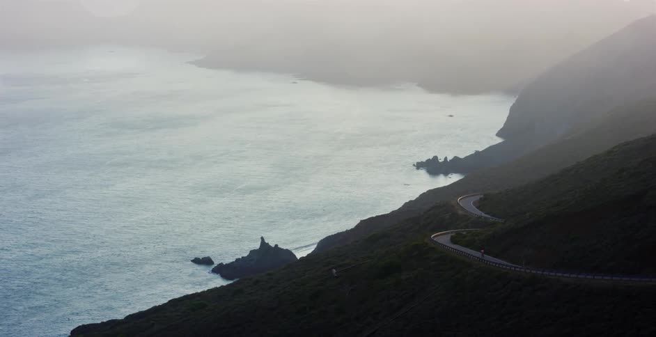 An aerial view shows a lone cyclist riding along a winding coastal highway through misty green hills at sunrise