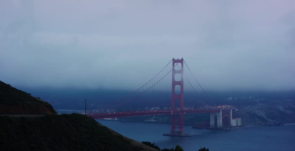 The iconic Golden Gate Bridge emerges from dense fog as traffic moves across during a moody, overcast evening