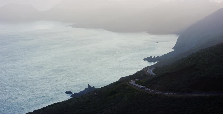 A single car navigates a winding coastal road along a steep, foggy mountainside overlooking the ocean