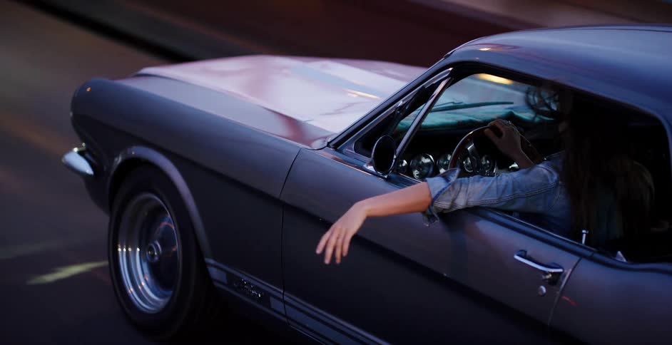 A carefree woman with long hair drives a classic grey muscle car at dusk, her hand playing in the wind outside the window