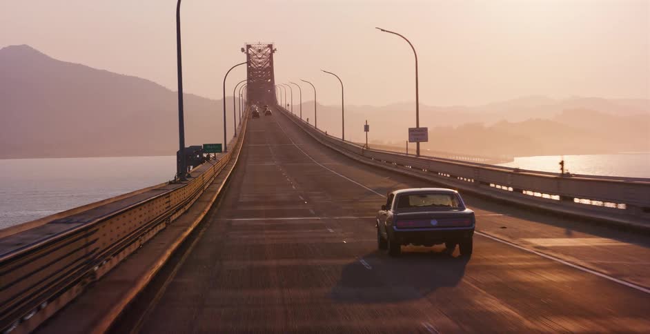 A classic black muscle car cruises across a long bridge spanning a wide bay during a hazy, golden sunset