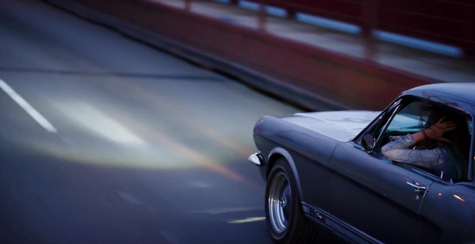 Close up of a woman driving her classic Ford Mustang fast on a bridge at dusk with her hair blowing in the wind