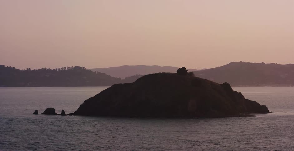 A silhouetted island floats in a calm bay against a hazy pink sky at dusk