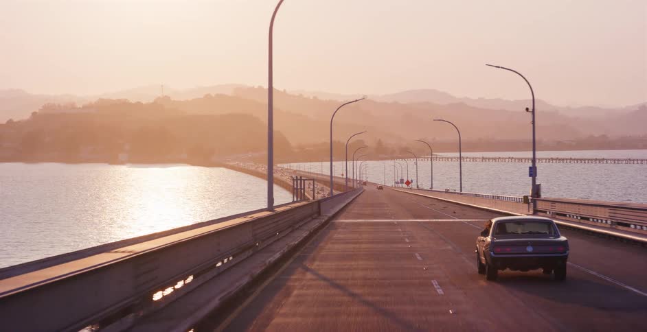 A vintage muscle car drives along a scenic coastal bridge over the water during a warm, golden sunset