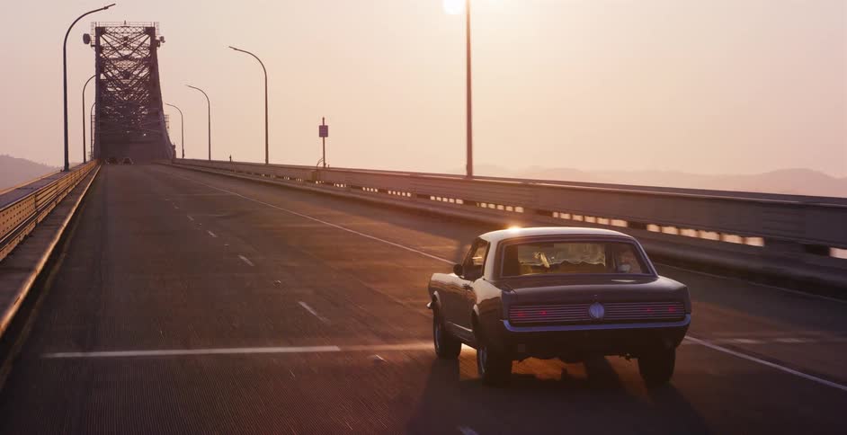 Rear view of a vintage black coupe driving along a long bridge during a beautiful hazy sunrise