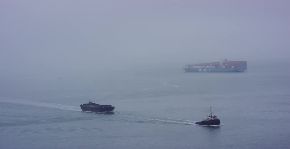 A tugboat pulls a barge across a foggy harbor with a large container ship visible in the background