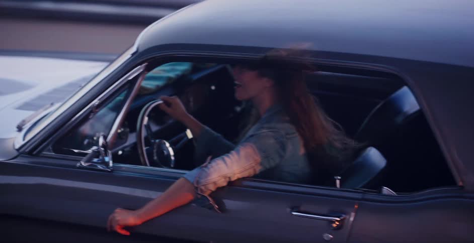 Happy woman with her hair blowing in the wind drives a classic muscle car with her arm out the window at dusk