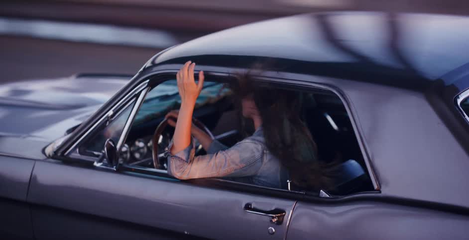 A free-spirited woman enjoys a drive in her classic silver Mustang, her long hair blowing in the wind as she cruises along at dusk