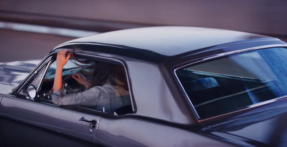 Slow motion shot of a woman with her hair blowing in the wind as she drives a vintage muscle car at dusk