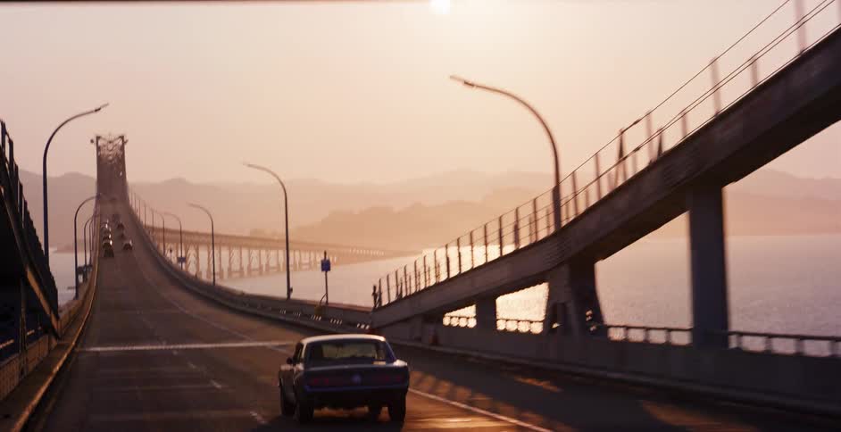 A cinematic reveal shot of a vintage muscle car driving across a bridge over the water at sunset