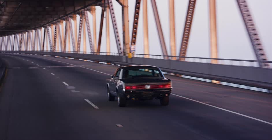 A classic black muscle car cruises along an empty industrial bridge as the warm light of sunset reflects off the steel structure