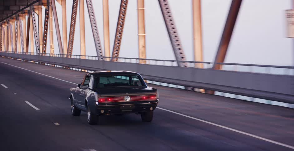 A classic black muscle car cruises across a long truss bridge during a golden sunset
