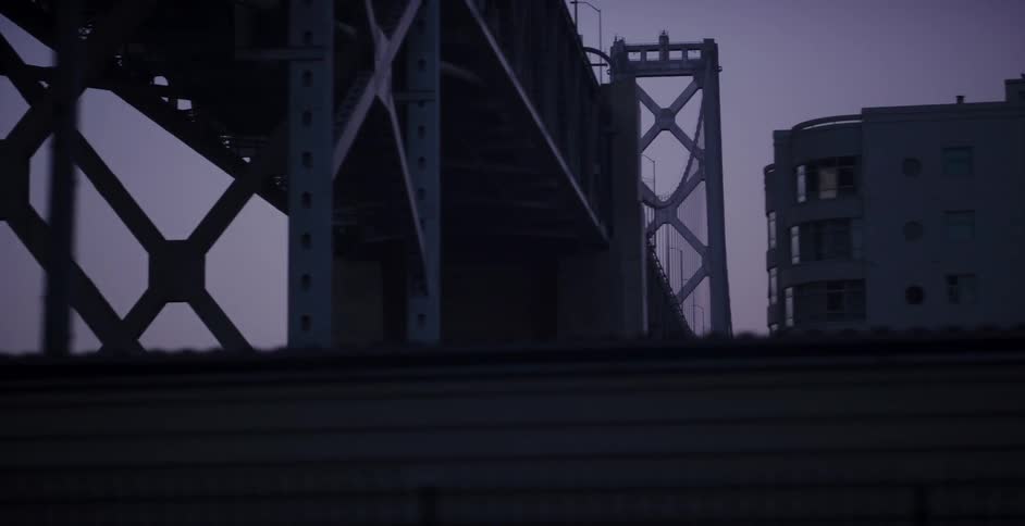A moody low-angle pan reveals the industrial steel structure of a city bridge at twilight