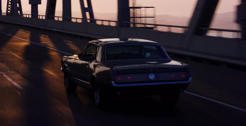 Following a classic black Mustang driving across a scenic bridge during a golden hour sunset