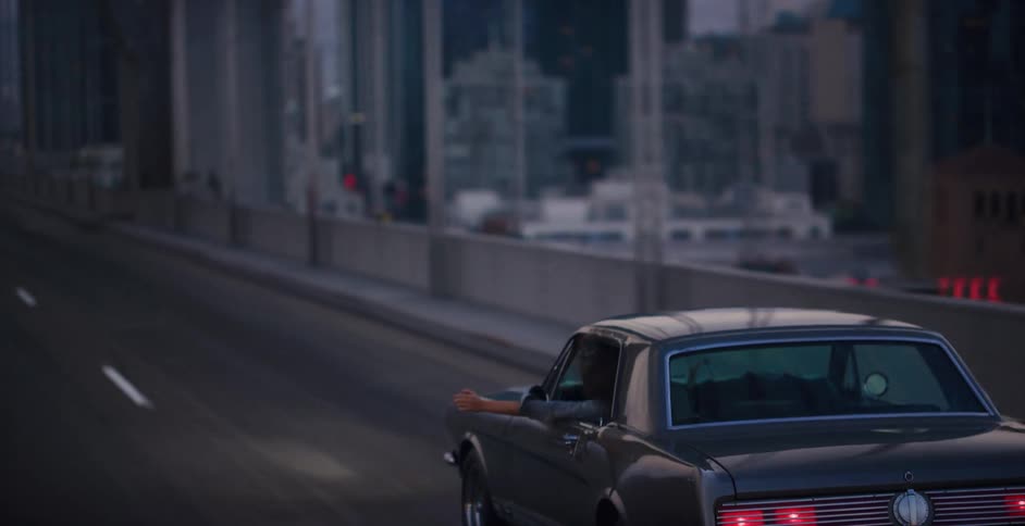 A person in a classic car enjoys a leisurely drive over a bridge with the city skyline blurred in the background at dusk
