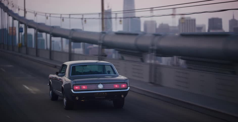 A classic muscle car drives over the Bay Bridge towards the San Francisco skyline at dusk