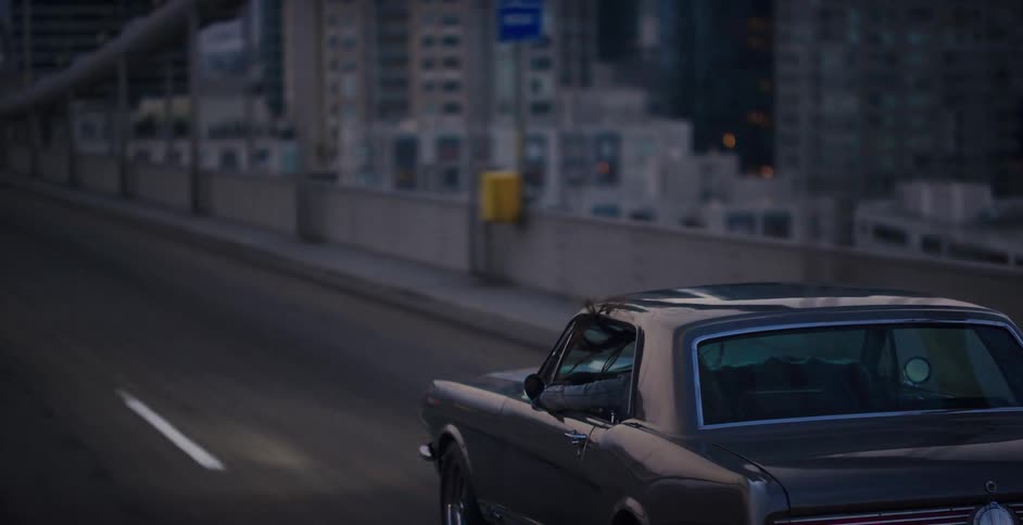 A woman cruises in a classic dark gray muscle car along a city bridge at dusk, with her hand out the window feeling the air