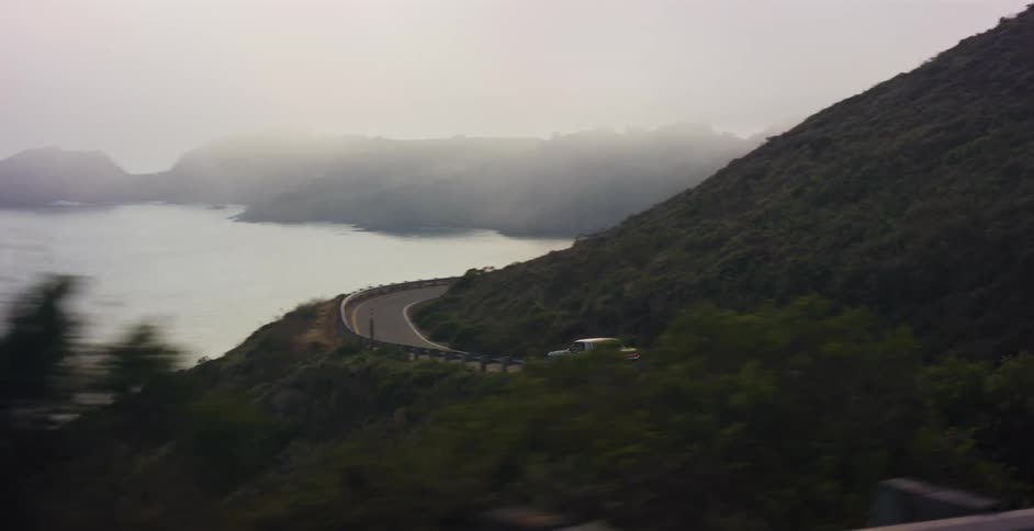 A point-of-view shot from a car driving along a winding coastal highway on a foggy day with the ocean below